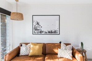 Un soggiorno moderno con un divano in pelle marrone, cuscini decorati, un quadro in bianco e nero che raffigura un paesaggio, una lampada a sospensione intrecciata e una piccola pianta in vaso su uno sgabello di legno.