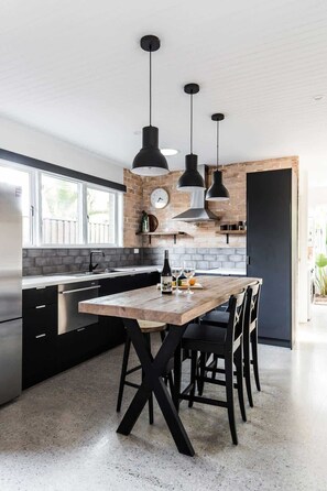 A modern kitchen with a wooden table, black chairs, hanging pendant lights, a clock, and a brick wall. - The Sunshine Bungalow Beautiful Noosa Home (Noosa)