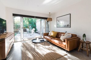 Ein modernes Wohnzimmer mit Ledersofa, Couchtisch und Fernseher. Eine Glastür öffnet sich zu einer Terrasse mit Tisch und Stühlen.