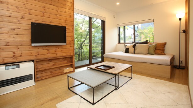 A modern living room with a flat-screen TV mounted on a wooden wall, a sofa with cushions, a coffee table, and a large window offering a view of trees. - KAMATA SANSO (Hakuba)