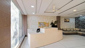 A reception area with a curved desk, a ceiling fan, patterned wallpaper, and a seating area with a sofa. - Hotel Shree Aaradhy (Ujjain)