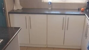 Une cuisine avec des meubles blancs, un plan de travail noir et une fenêtre équipée de stores.