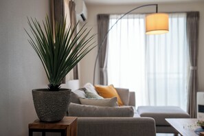 A modern living room with a potted plant on a wooden side table, a grey sofa with cushions, and a large window with curtains. - LABRADORITE (Sapporo)
