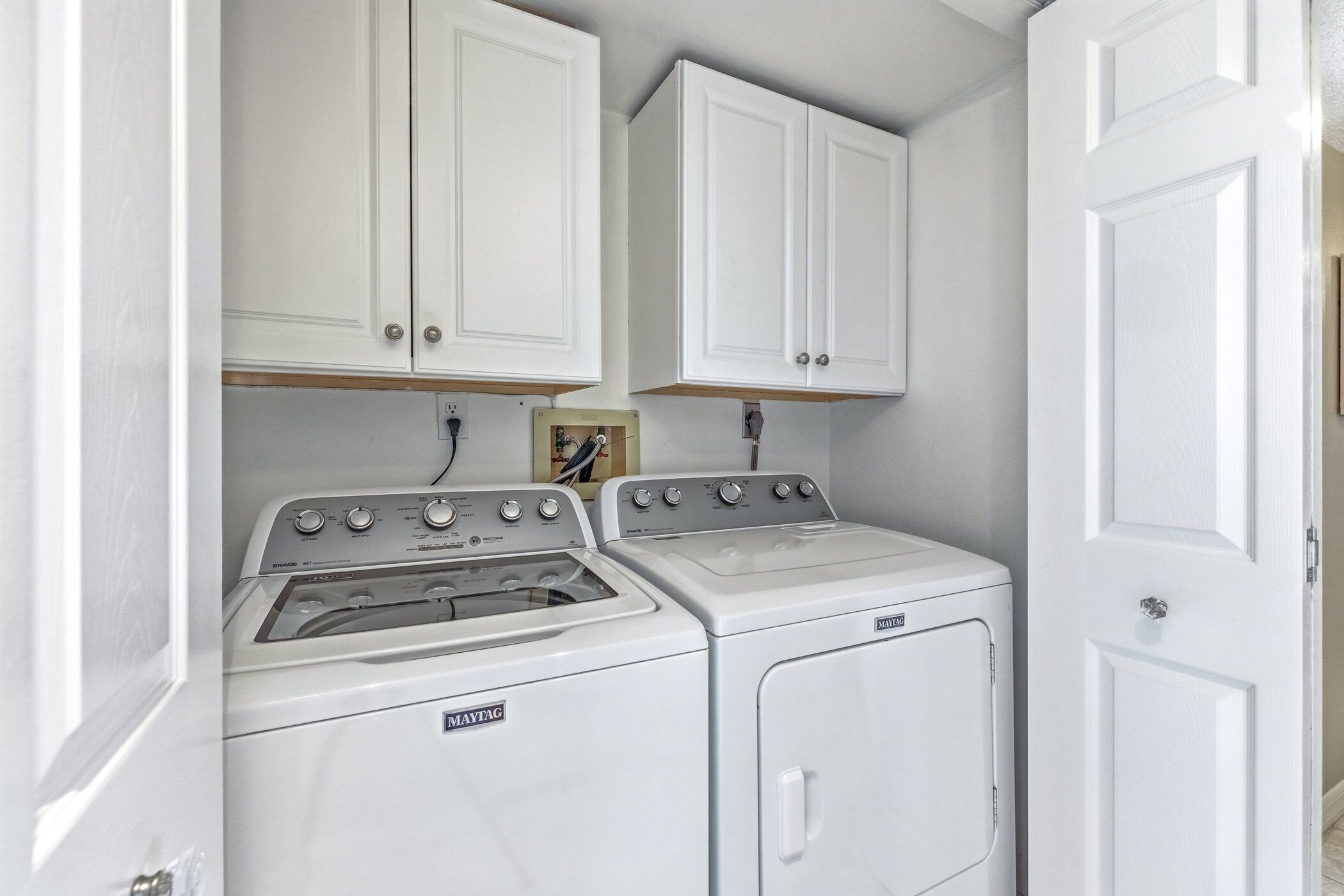 A laundry room with a front-loading washer and dryer, white cabinets above, and a door to the right.
