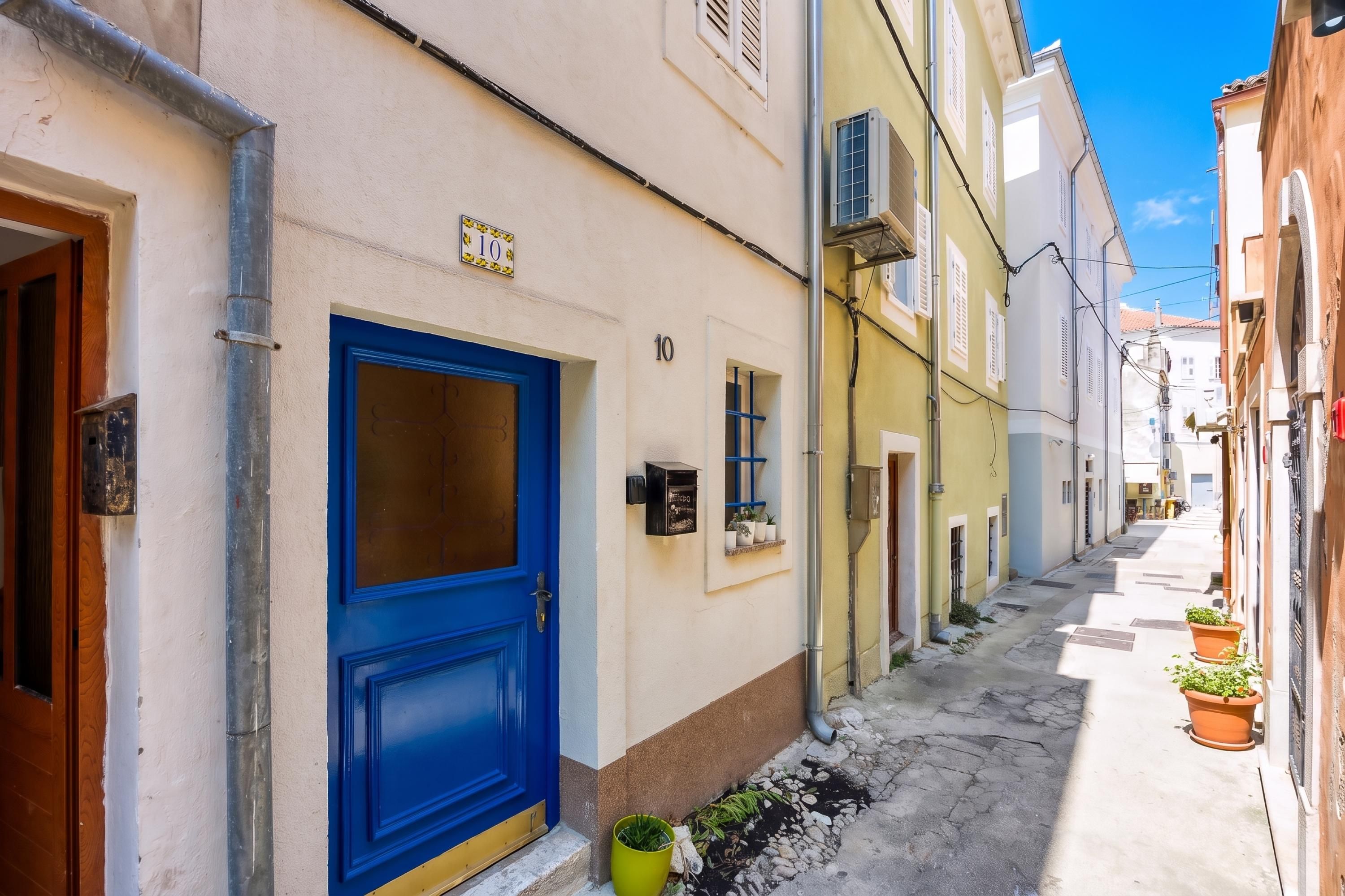 A narrow street with buildings on either side, a blue door, and a small green pot on the ground.