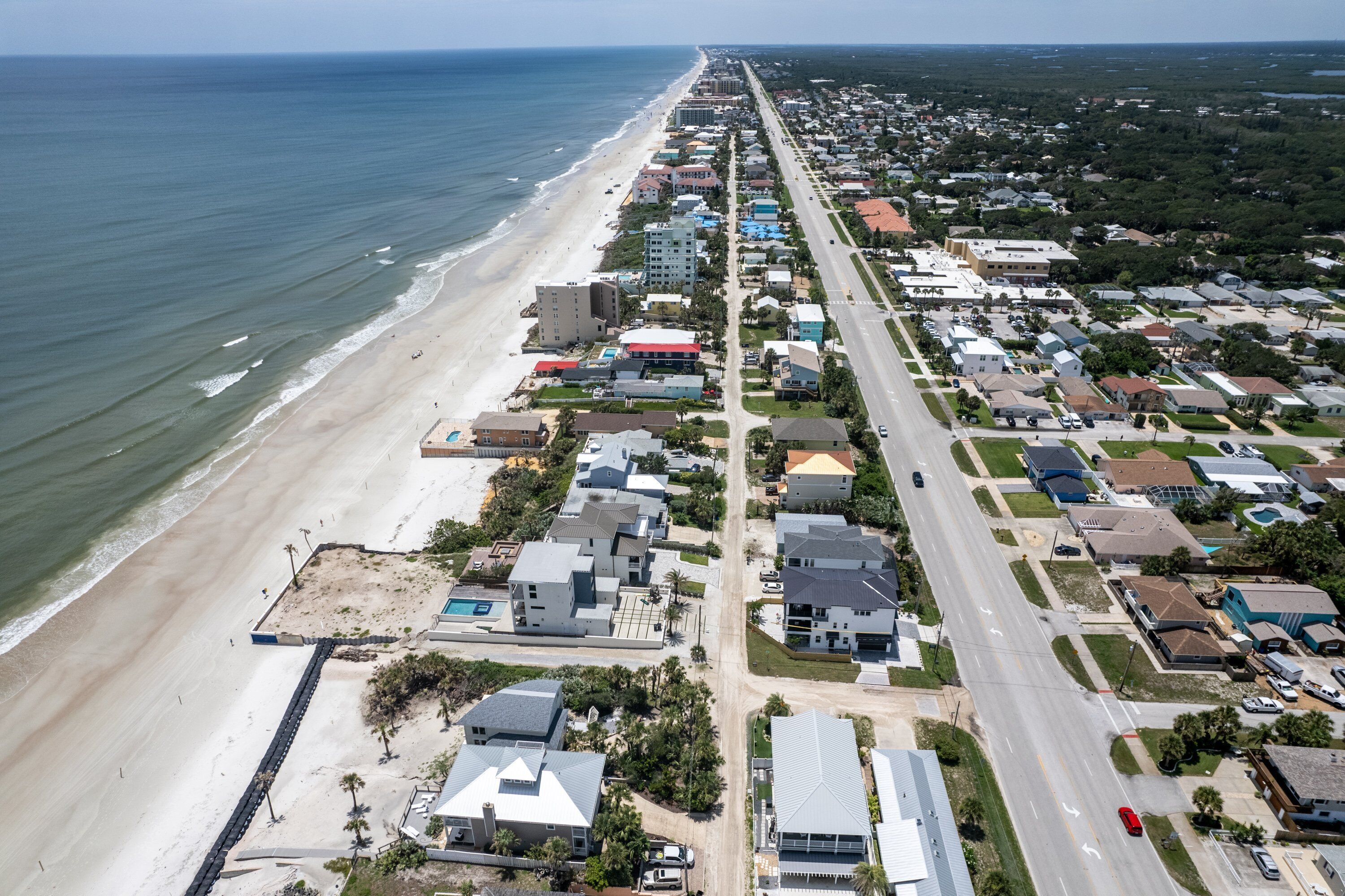 An aerial view of a coastal residential area with a beach, houses, and a road.