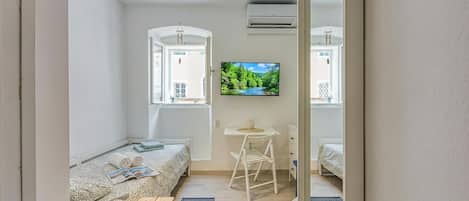 A compact bedroom with a bed, a small desk, a chair, and a TV on the wall.