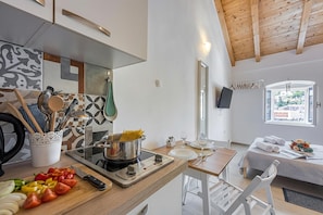 Una cocina moderna con estufa, tabla de cortar y utensilios. Se ve un comedor con mesa y sillas.