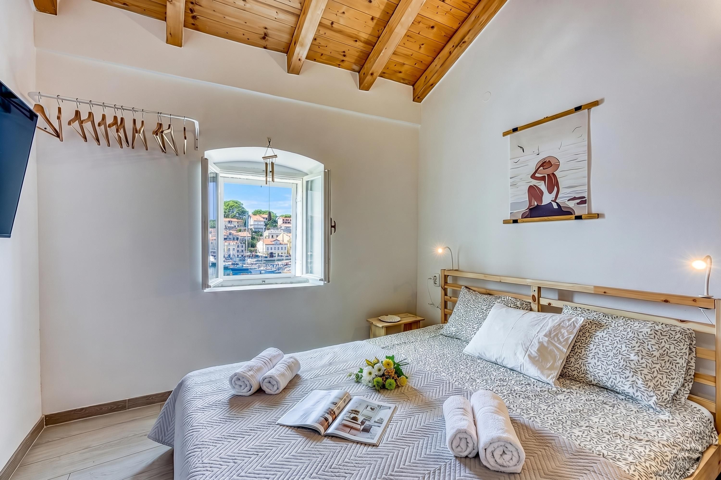 A bedroom with a bed, a window with a view of buildings and a marina, a wooden ceiling, and a wall-mounted artwork.