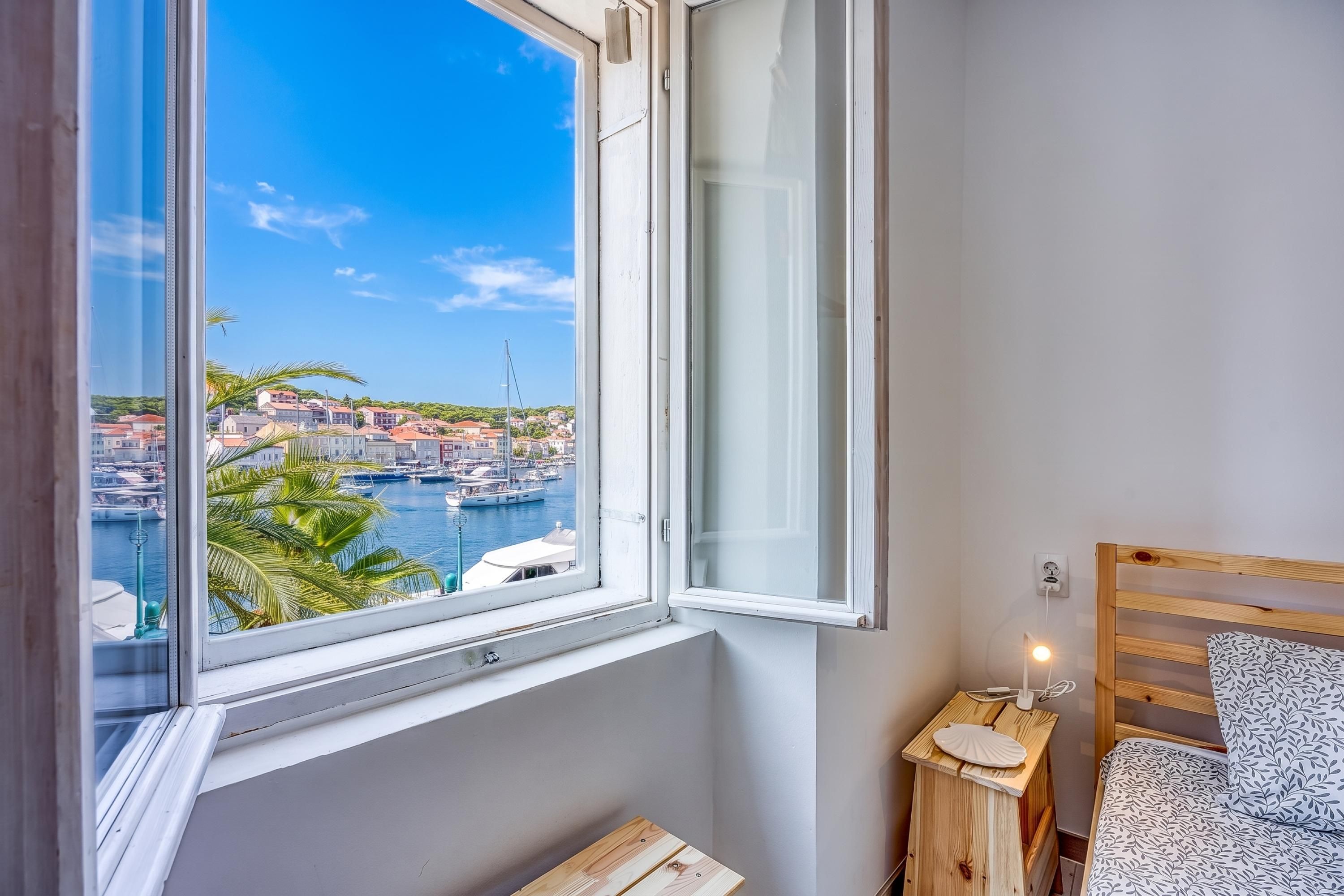 A bedroom with a view of a harbor, a wooden bedside table with a lamp, and a bed with a patterned pillow.