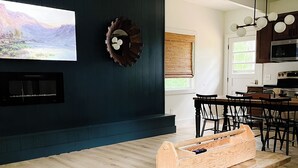 Una sala de estar moderna con una pared de acento oscura, un televisor de pantalla plana, un banco de madera y un comedor con sillas y una mesa.