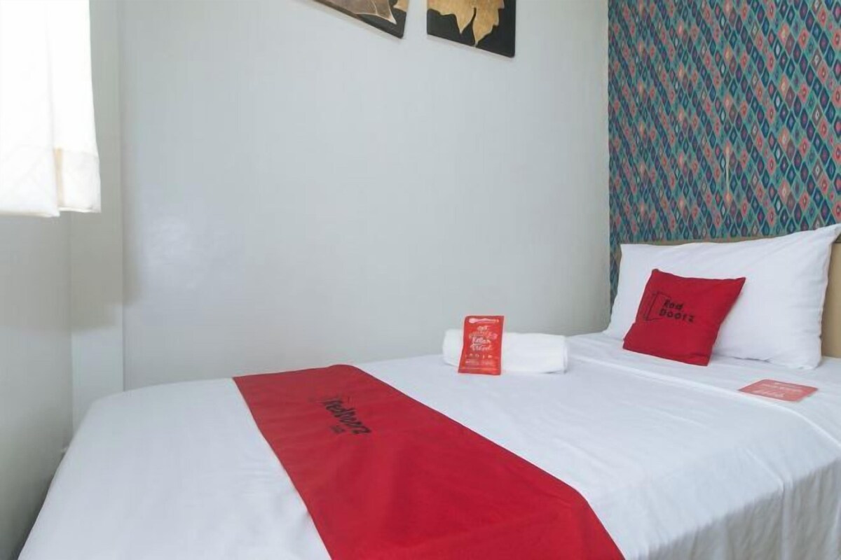 Una cama bien hecha con una colcha roja, almohadas blancas y una almohada roja con texto. Hay un pequeño objeto rojo sobre la cama, quizás un libro o una tarjeta. La habitación tiene una pared blanca lisa y una cortina con estampado.