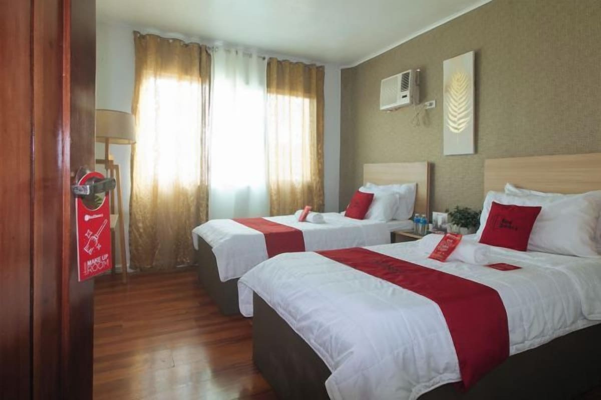 A hotel room with two beds, a wooden floor, a window with curtains, and a wall-mounted air conditioner.