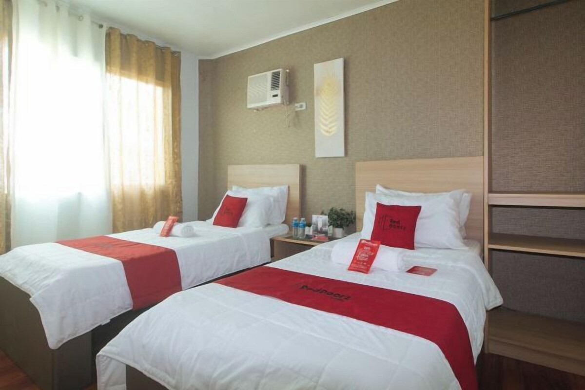 Habitación de hotel con dos camas, cada una con ropa de cama blanca y detalles en rojo, una mesita de noche con artículos de aseo, y un aire acondicionado de pared.