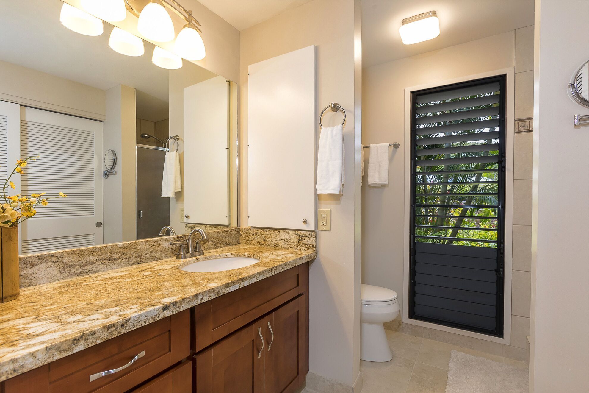 Une salle de bain dotée d’un comptoir en granit, d’un grand miroir, de toilettes avec une fenêtre et d’un espace douche.