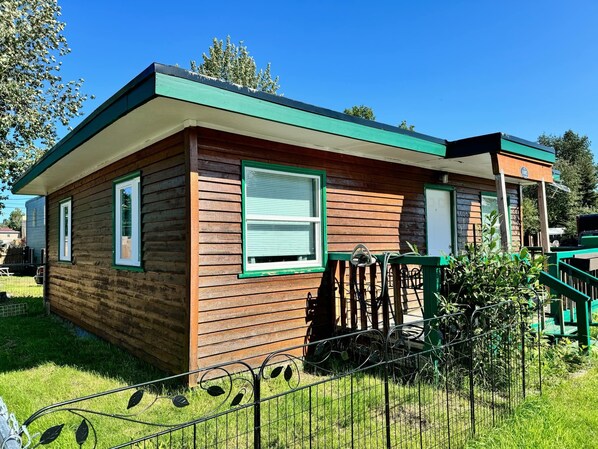 A small wooden cabin with a green roof, a fenced yard, and a small porch.