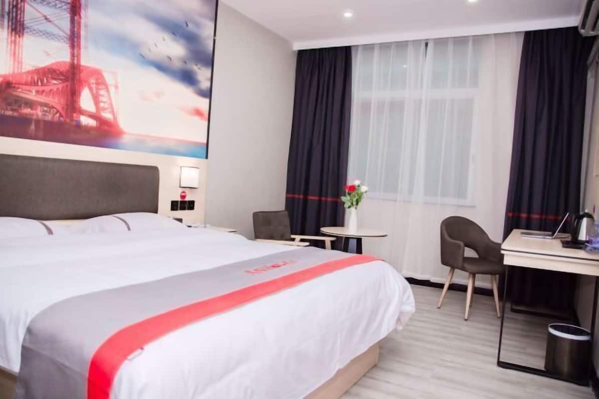 Quarto de hotel com uma cama, escrivaninha, cadeira, mesa pequena e janela com cortinas.