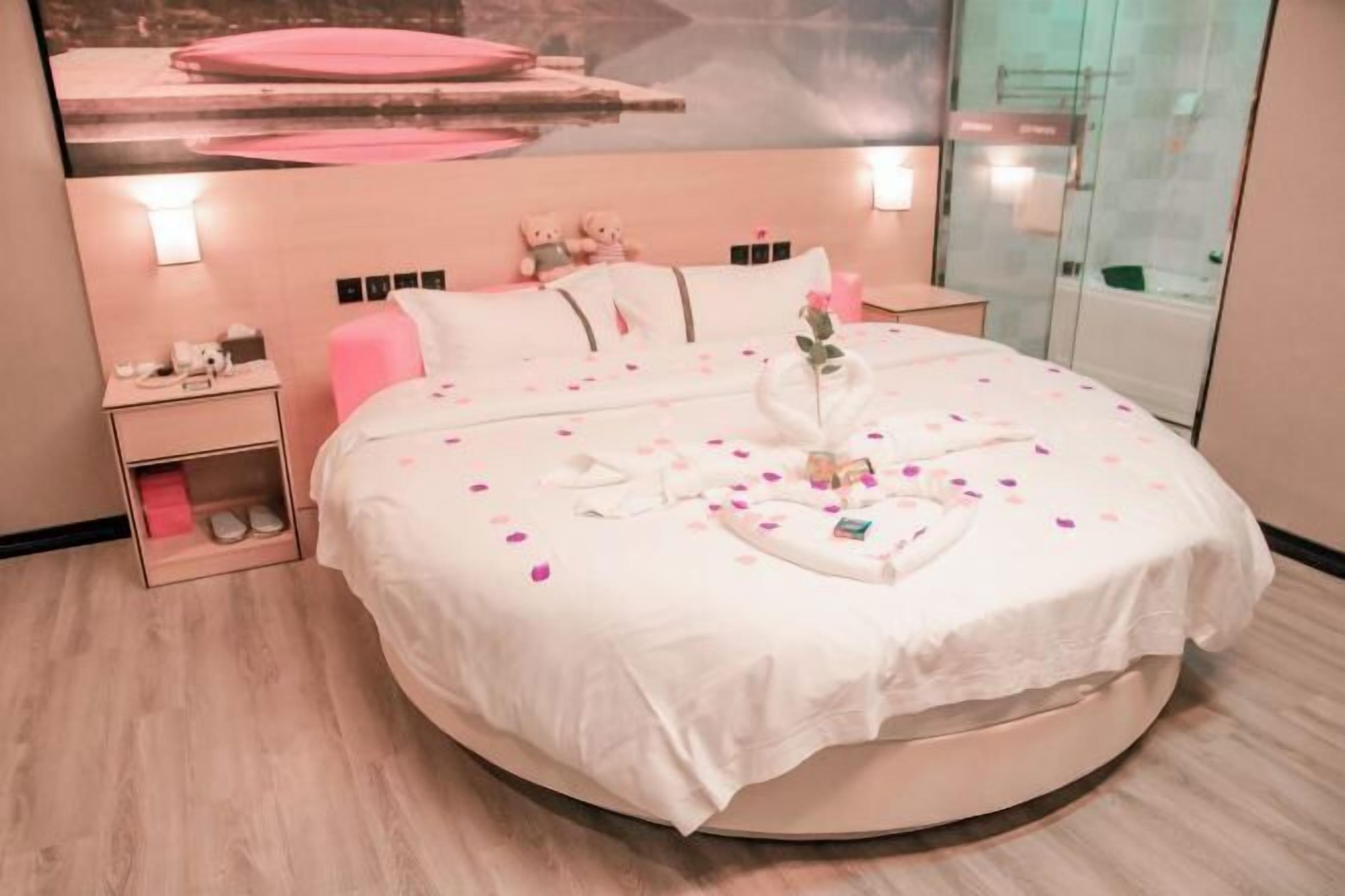 A round bed with a white bedspread, pillows, and a bouquet of flowers. There is a nightstand with a lamp and a small table with a teddy bear. A poster is on the wall above the bed.