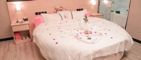 A round bed with a white bedspread, pillows, and a bouquet of flowers. There is a nightstand with a lamp and a small table with a teddy bear. A poster is on the wall above the bed.