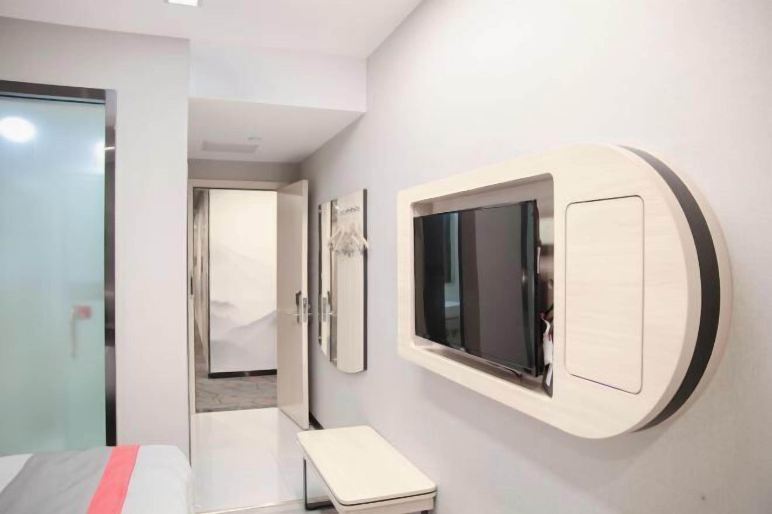 A modern hotel room with a bed, a small bedside table, a wall-mounted TV, and a door leading to another room.