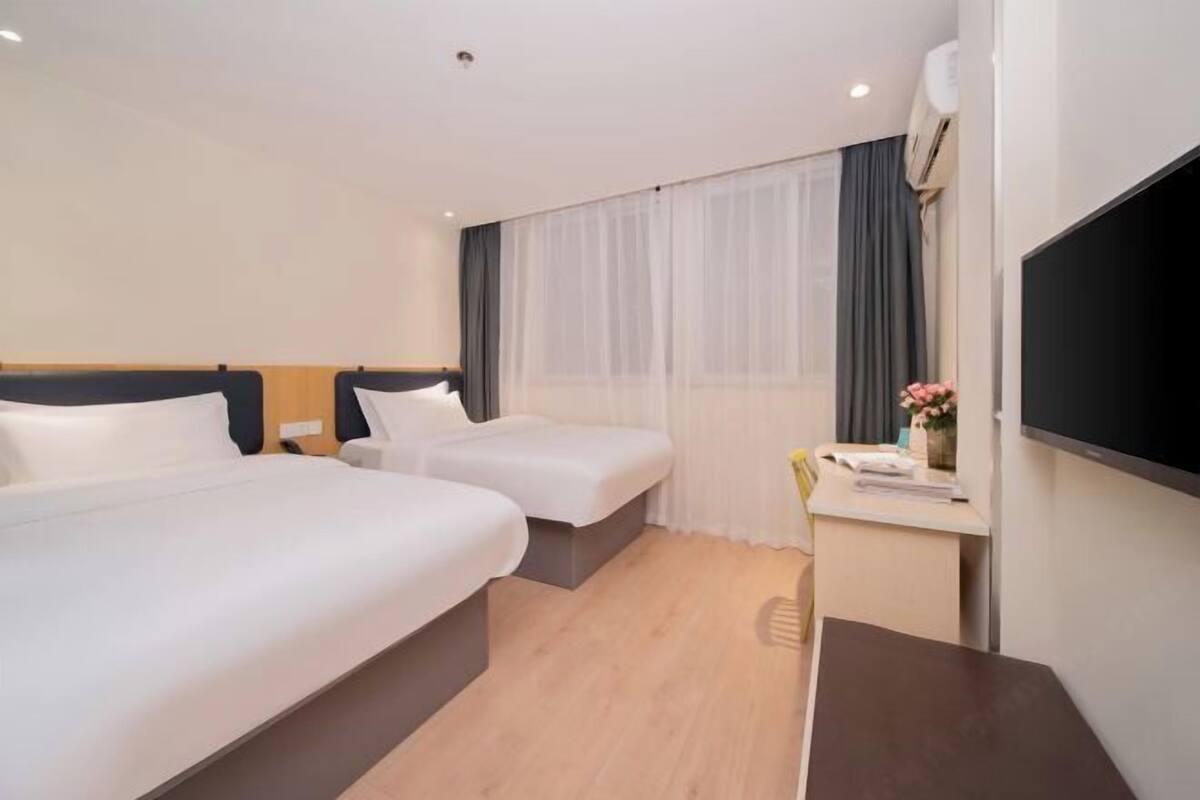 A hotel room with two beds, a desk, a flat-screen TV, and a large window with sheer curtains.