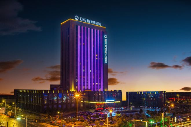 A tall hotel building with illuminated vertical lines, a parking lot, and street lights.