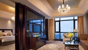 Habitación de hotel con una cama grande, una zona de estar con una mesita de café, televisión y vistas a la ciudad a través de amplios ventanales.