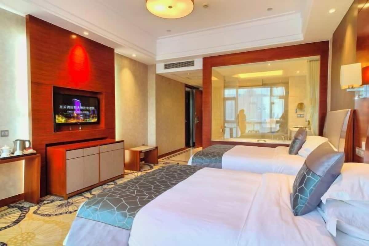 A hotel room with two beds, a TV, a desk, and a bathroom visible through a glass door.