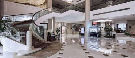 A spacious lobby with a central staircase, a reception desk, and a modern chandelier.
