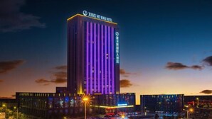 A tall hotel building with vertical purple lighting, a sign reading "XING HE WAN HOTEL," and a parking lot with cars. - Xinghewan International Hotel (Sheyang)