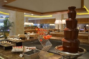 Zona de buffet con una cascada de chocolate como pieza central, diferentes estaciones de comida y una decoración interior moderna.