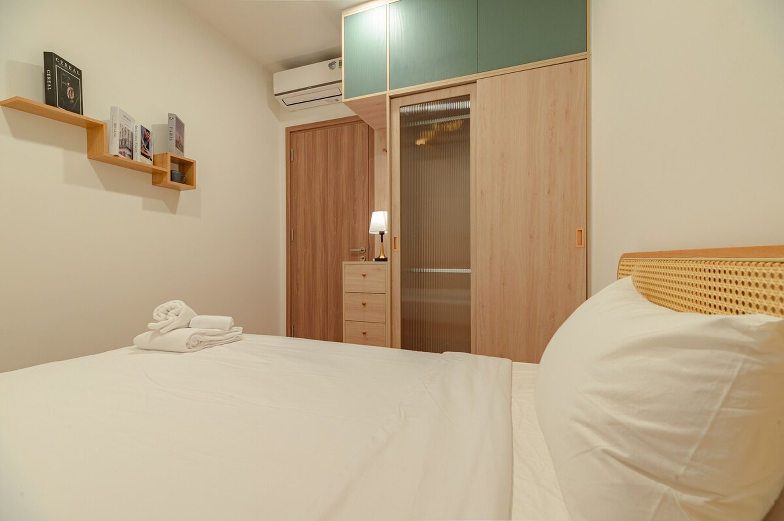Ein ordentlich bezogenes Bett mit einem gefalteten Handtuch, ein Holzregal mit Büchern, ein Kleiderschrank und eine Nachttischlampe.