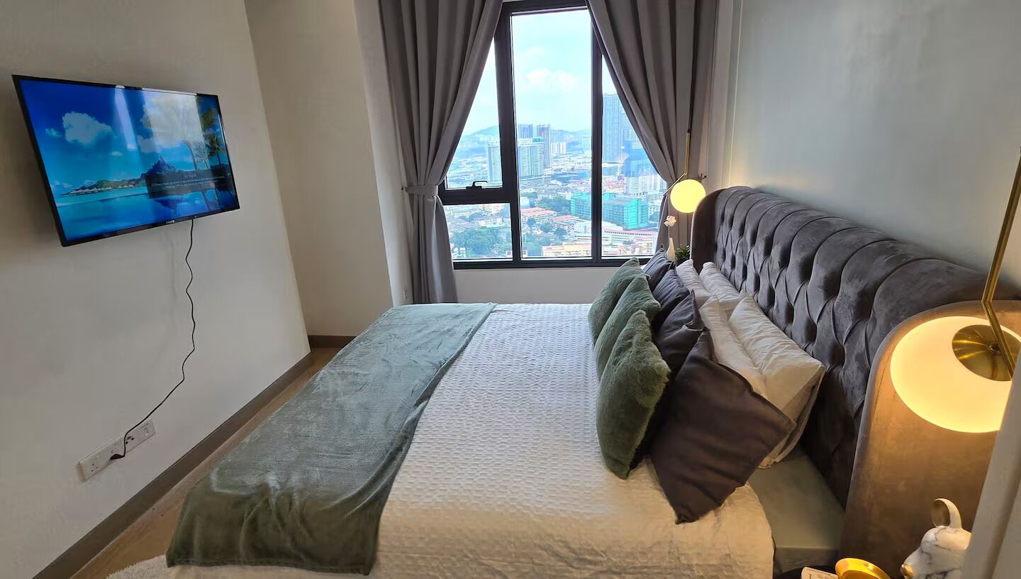 Un dormitorio con cama, televisor montado en la pared, ventana con vista a la ciudad y lámpara de noche.