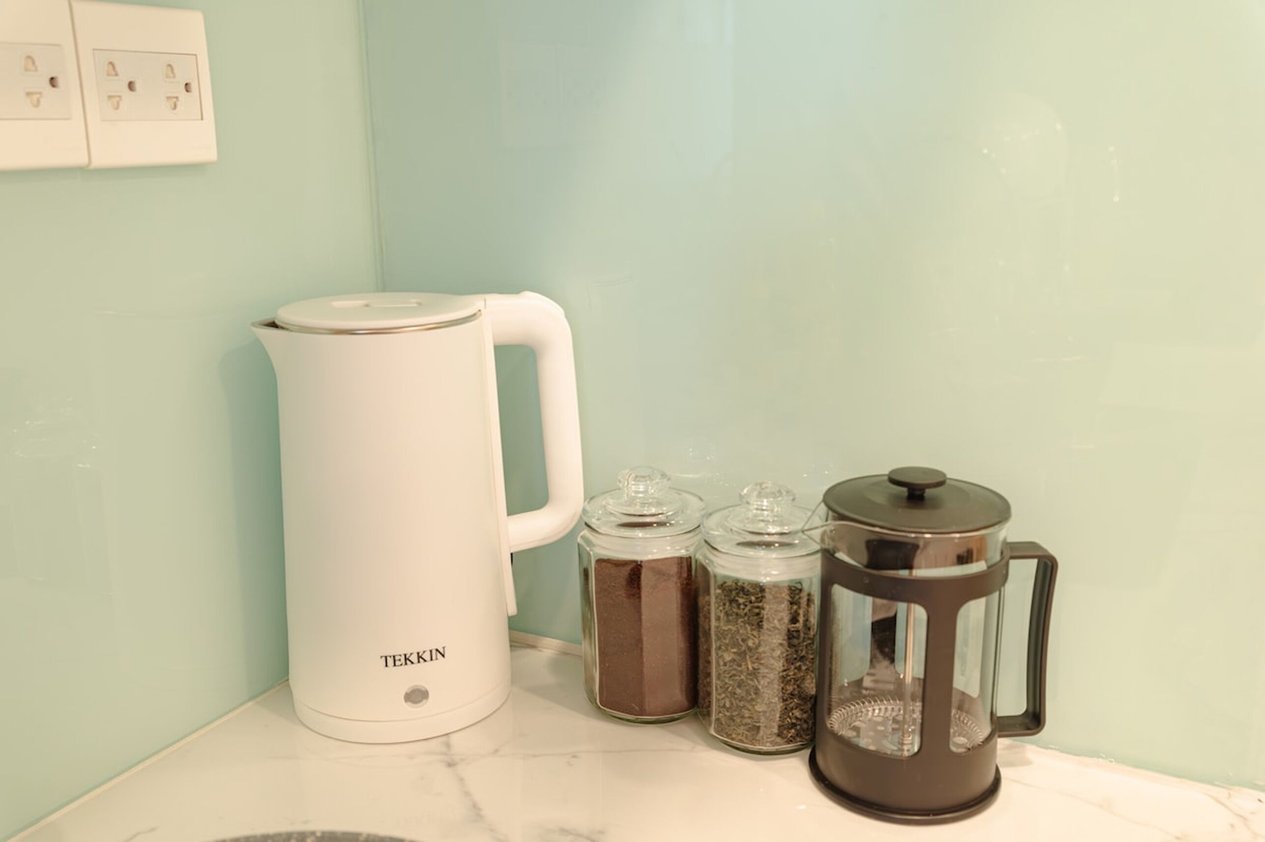 Una cafetera blanca, dos recipientes de vidrio con café molido y una prensa francesa sobre una encimera de mármol.