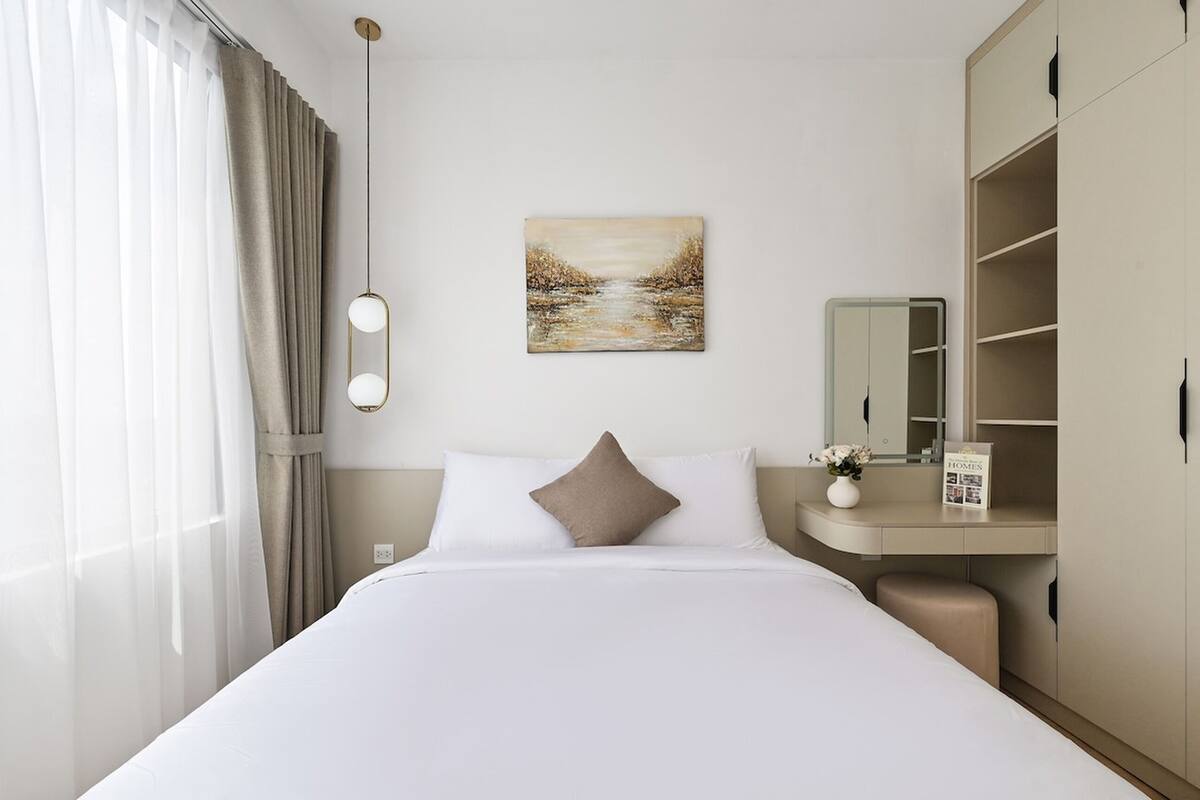A hotel room with a bed, a bedside table with a vase, a painting, a wardrobe, and a window with curtains.