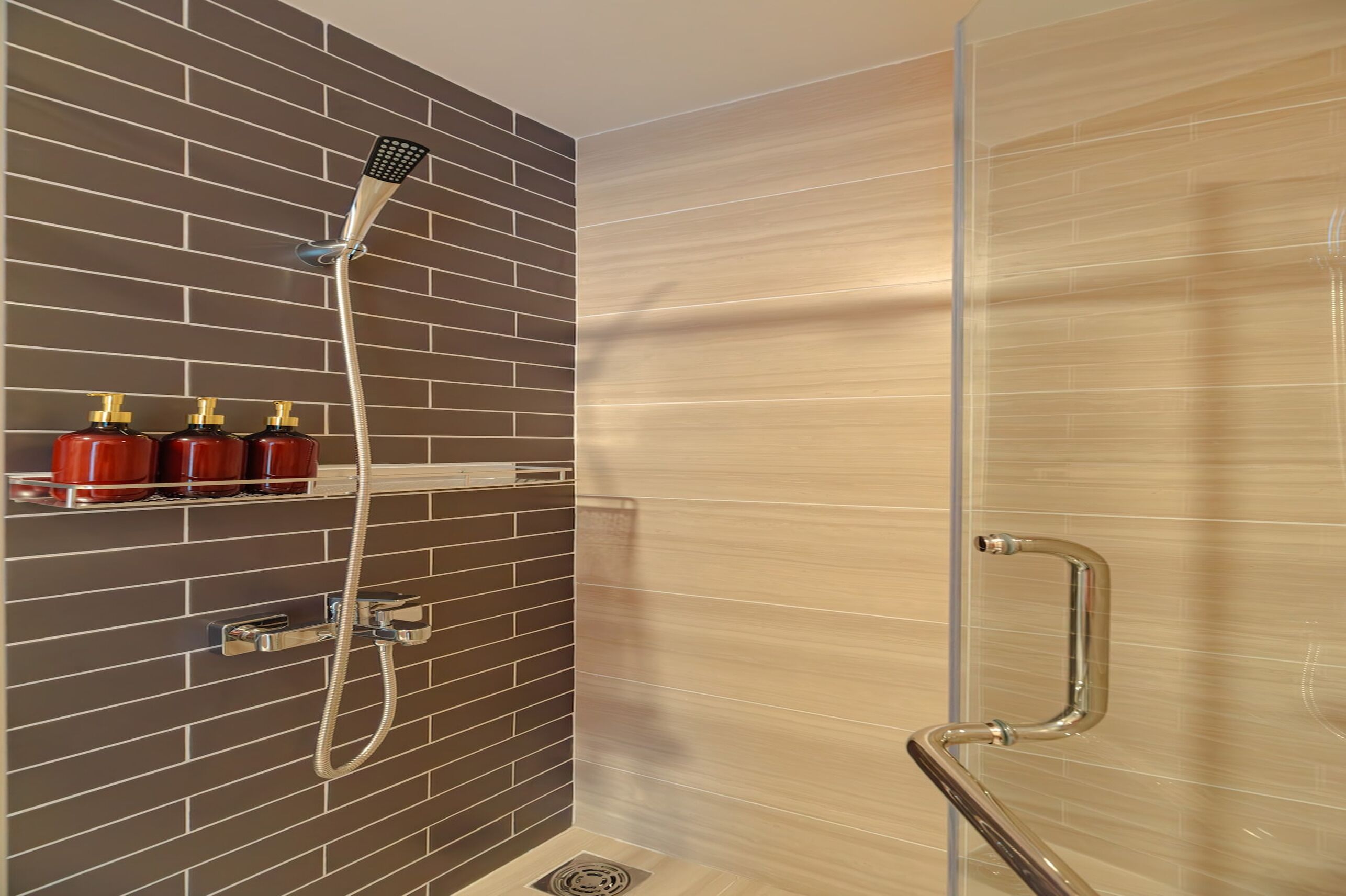 A modern bathroom with a shower, a handrail, and a shelf with three bottles.