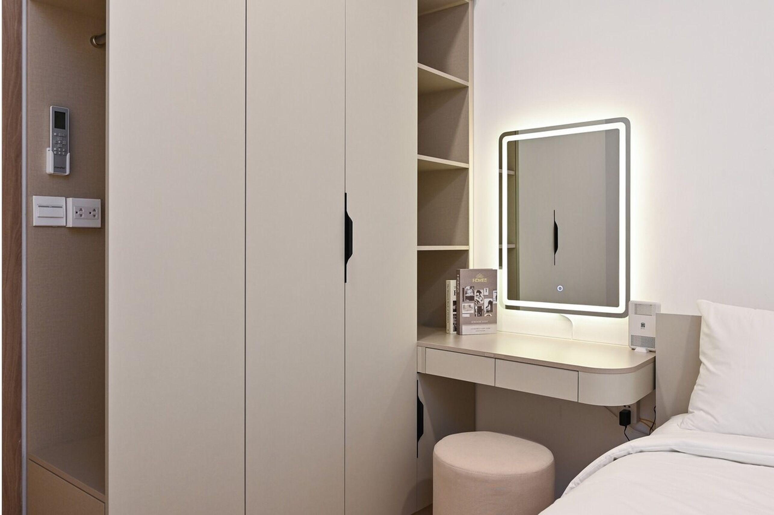 A hotel room with a bed, a bedside table with a lamp, a mirror, a wardrobe, and a shelf with a book.