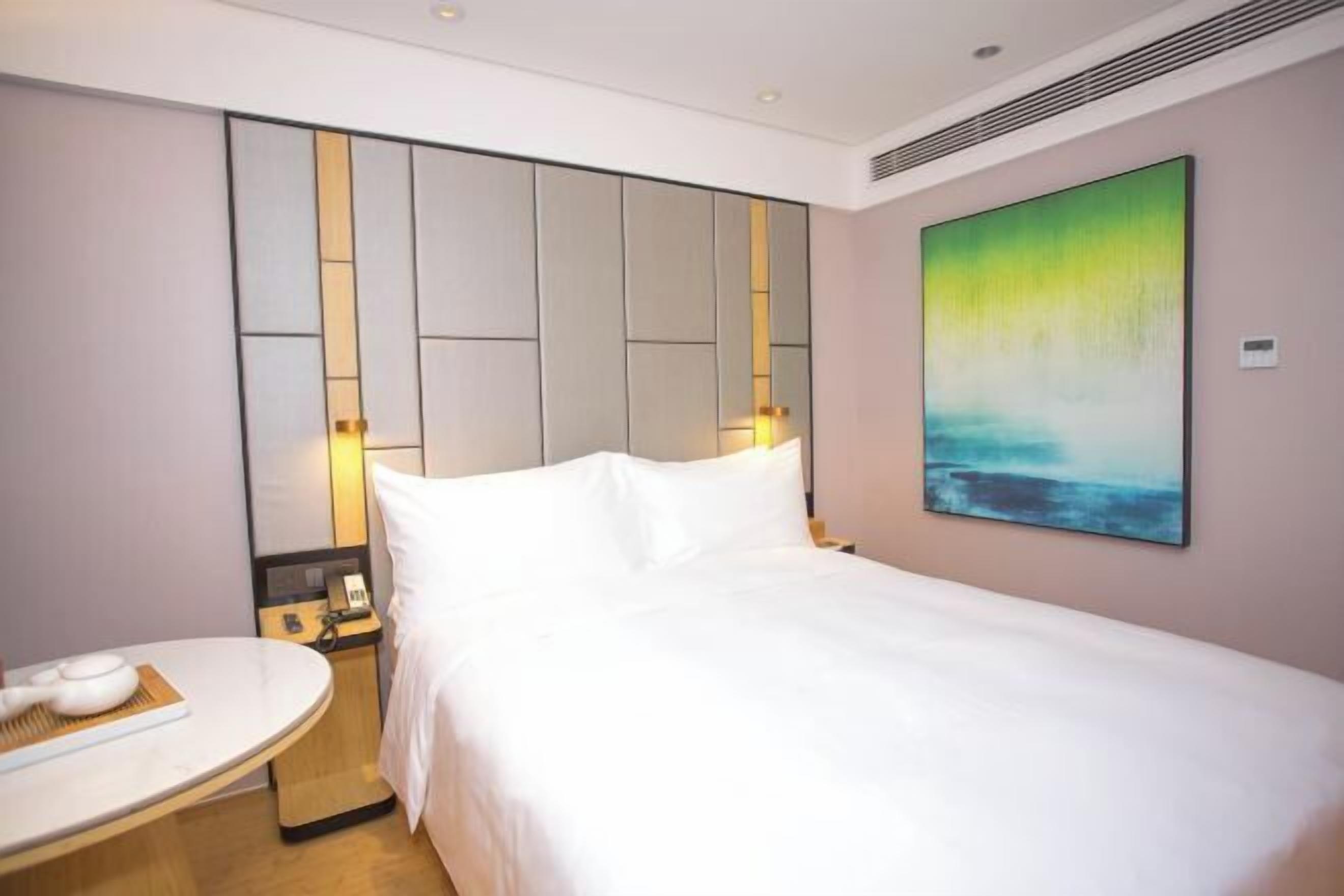 A modern hotel room with a large bed, a bedside table with a lamp, and a painting on the wall.