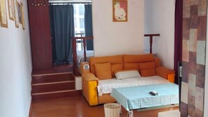 A room with a sofa bed, a small table with a tablecloth, a chair, a waste basket, and a staircase leading to another room. - Hanlin Gangwan Apartment (Zhuhai)