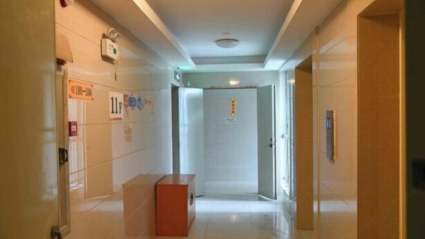 A hotel corridor with a reception desk, a door with a sign, and a wall-mounted emergency light. - Hanlin Gangwan Apartment (Zhuhai)