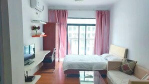 A small, well-lit room with a bed, a sofa, a TV, and a window with curtains. - Hanlin Gangwan Apartment (Zhuhai)