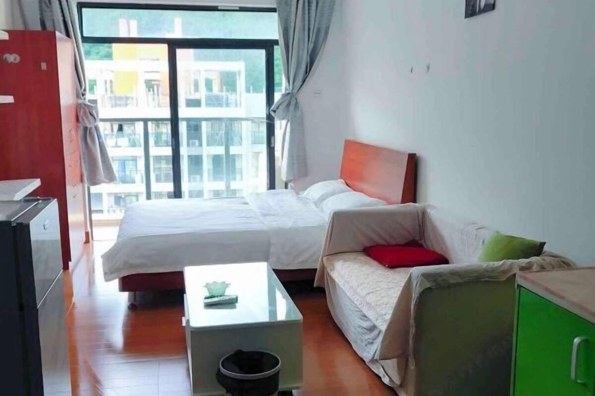 A room with a bed, a sofa, a small table, a green cabinet, and a window with a view of buildings.
