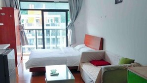 A room with a bed, a sofa, a small table, a green cabinet, and a window with a view of buildings. - Hanlin Gangwan Apartment (Zhuhai)