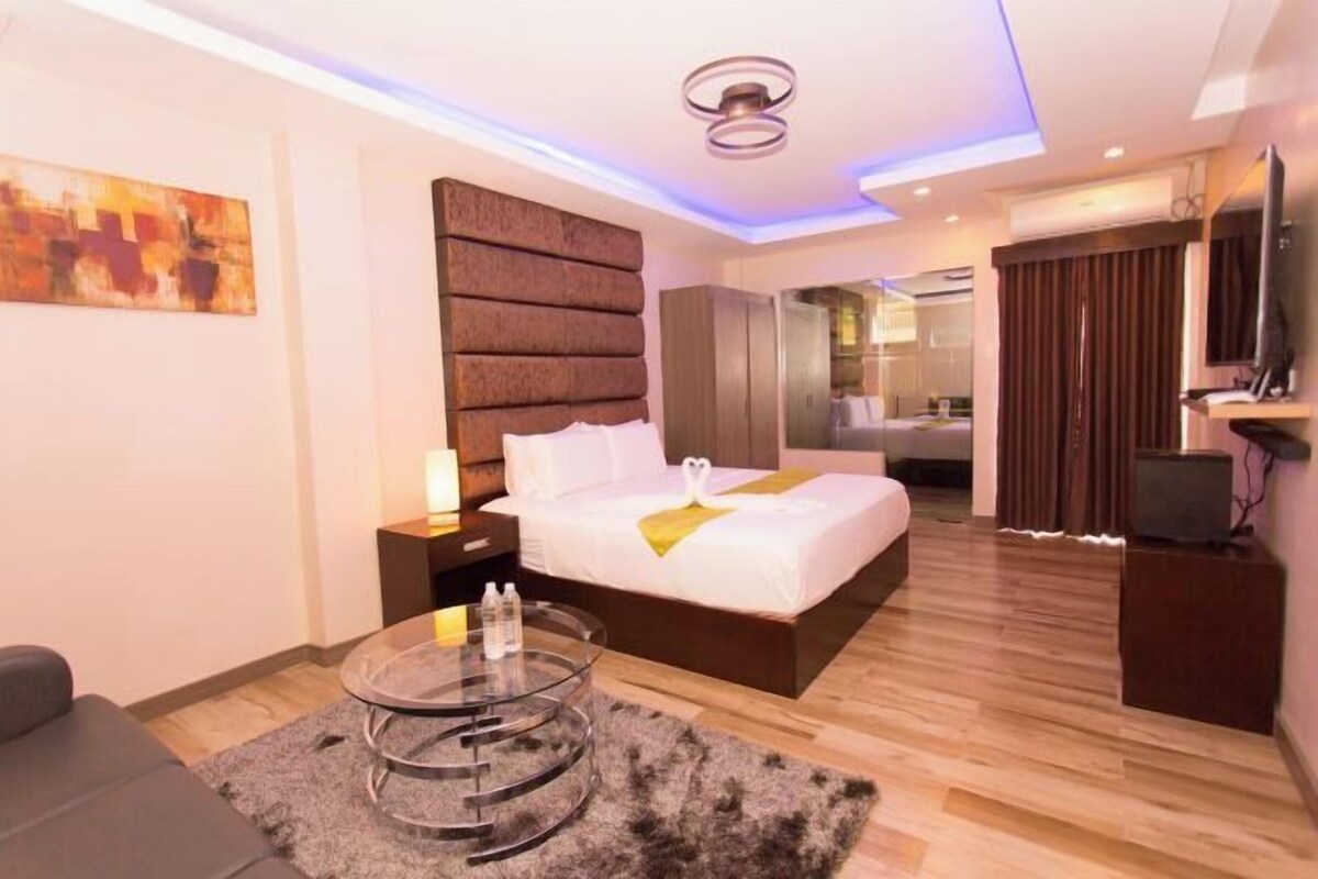 A modern hotel room with a large bed, a glass coffee table, a TV, and a painting on the wall.