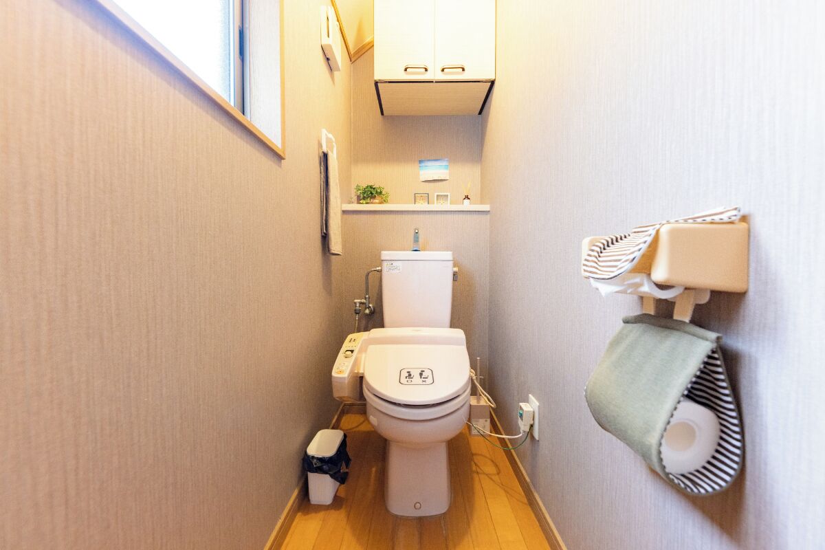 A compact bathroom with a toilet, a small trash bin, a towel dispenser, and a shelf with a plant and a picture.