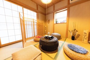 Una habitación japonesa tradicional con suelo de tatami, una mesa baja de tatami, un taburete redondo tejido y un pequeño estante de madera con adornos.