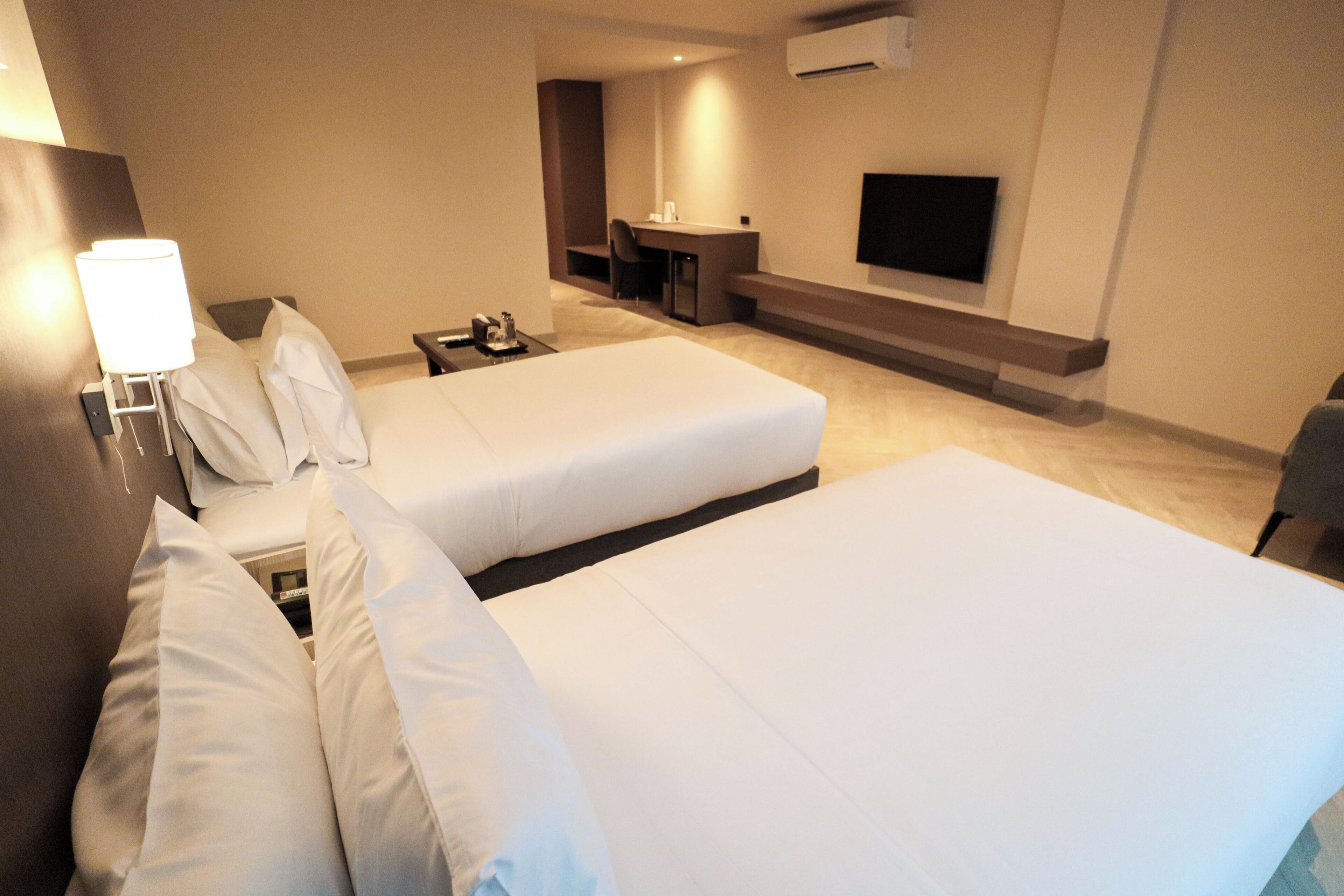 A hotel room with two beds, a desk, a chair, a TV, and a lamp.