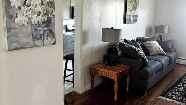 A living room with a dark wood floor, a grey sofa with patterned cushions, a small wooden side table, and a painting of white flowers on the wall. - Welcome to The Tennerton House-Charming 2-bedroom with AC and WiFi in Buckhannon (Buckhannon)