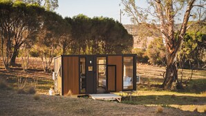 A small cabin with a wooden exterior, a visible bedroom with a bed and window, and a patio area with a table and chairs. - Rowsley Valley Tiny House 1 by Tiny Away (Glenmore)