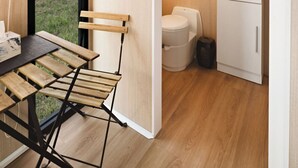 A compact room with a wooden floor, a small table with a chair, a window with a view of the outdoors, and a compact kitchenette with a sink and a mirror. - Rowsley Valley Tiny House 1 by Tiny Away (Glenmore)